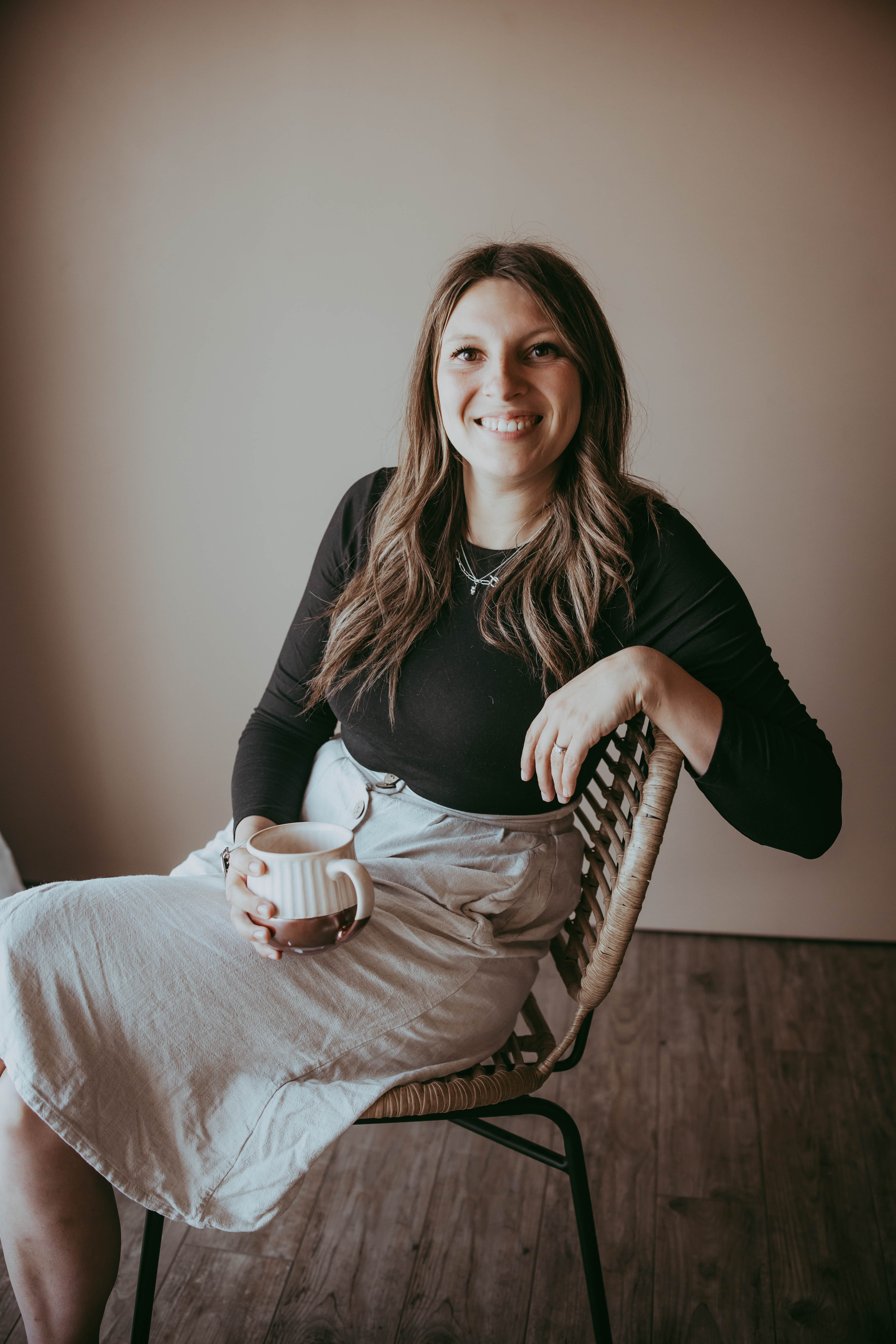Friendly, smiling woman with long brown hair sitting in a wicker chair and holding a mug in a warm, professional setting.