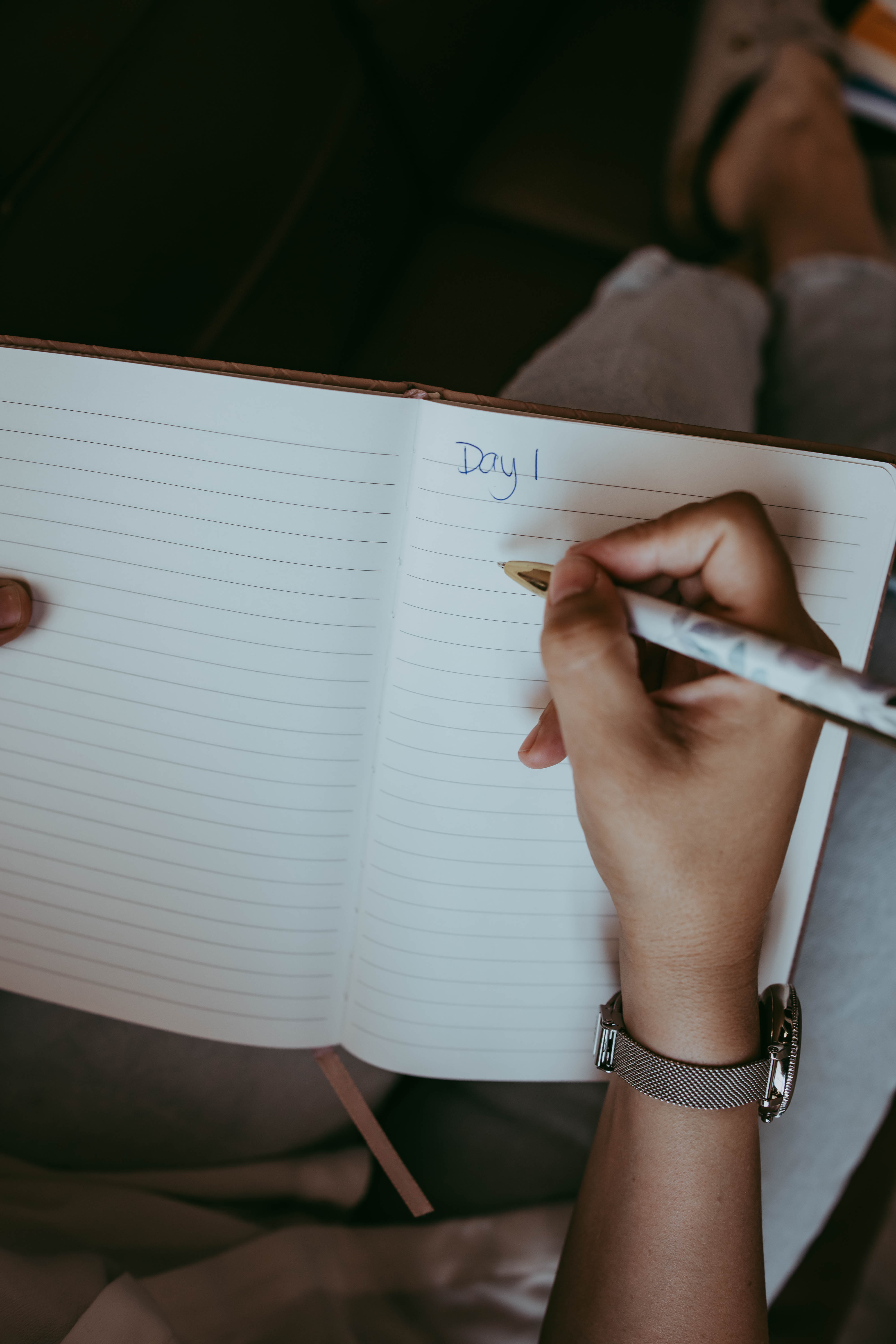 A person wearing a silver watch writes "Day 1" in a notebook, beginning a self-reflection or couples counseling journey.